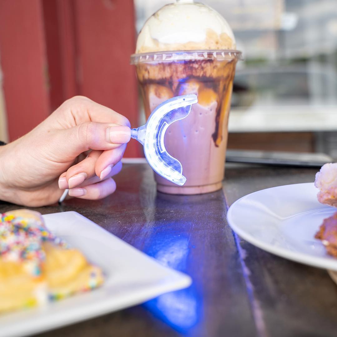 Woman holding NovaSmile Premium Teeth Whitening kit led mouthpiece in restaurant next to her chocolate milkshake, waffle and doughnuts